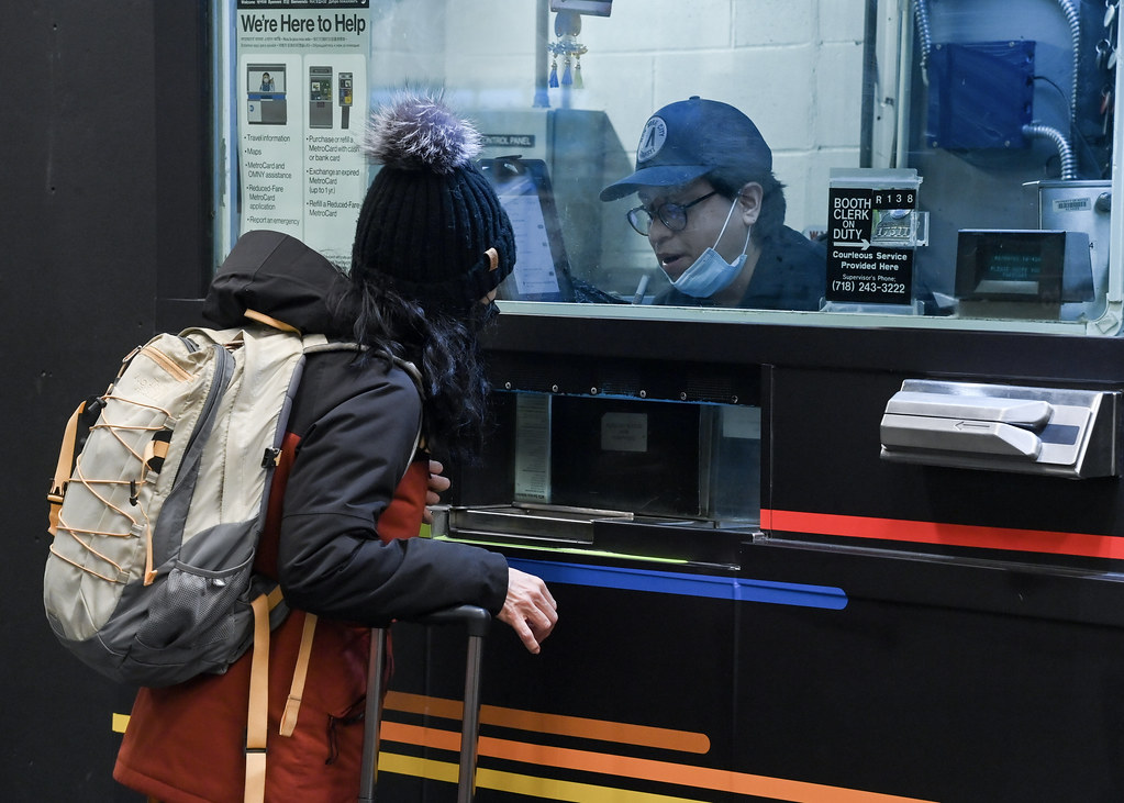 A traveller with a backpack speaks to a staff member through the window of a subway customer service booth.