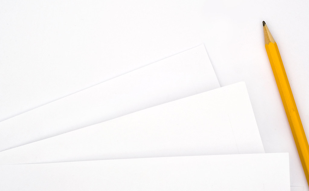 A pencil lies beside overlapping blank sheets of paper on a pale desk.