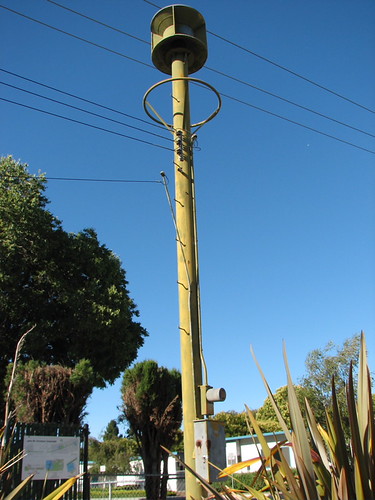 A cylindrical emergency siren mounted at the top of a tall wooden pole, with overhead cables and trees against a blue sky.