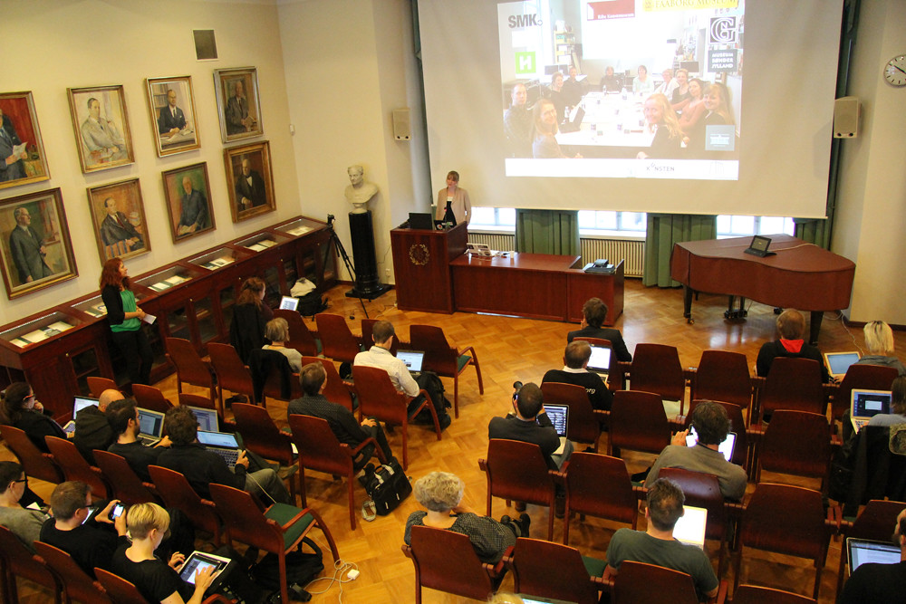 A speaker presents to seated participants using laptops in a hall with framed portraits and a projection screen.