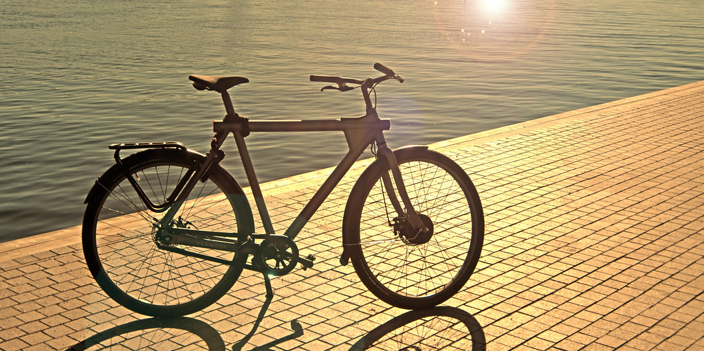 A black electric bicycle parked beside the water in low sunlight.