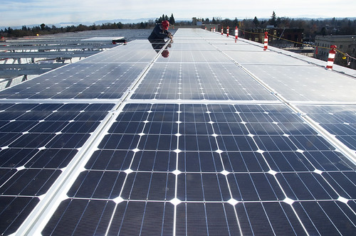Rows of solar panels installed across a flat roof, with a worker in the distance.