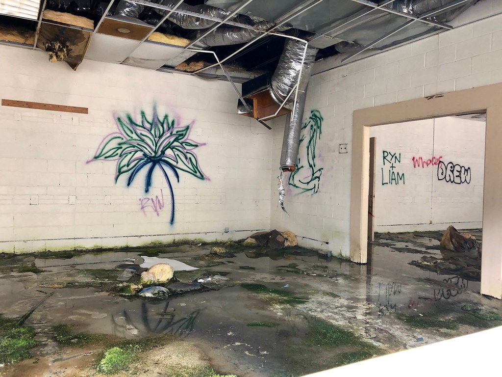 An abandoned classroom with standing water, mould, missing ceiling panels and hanging ventilation ducts.
