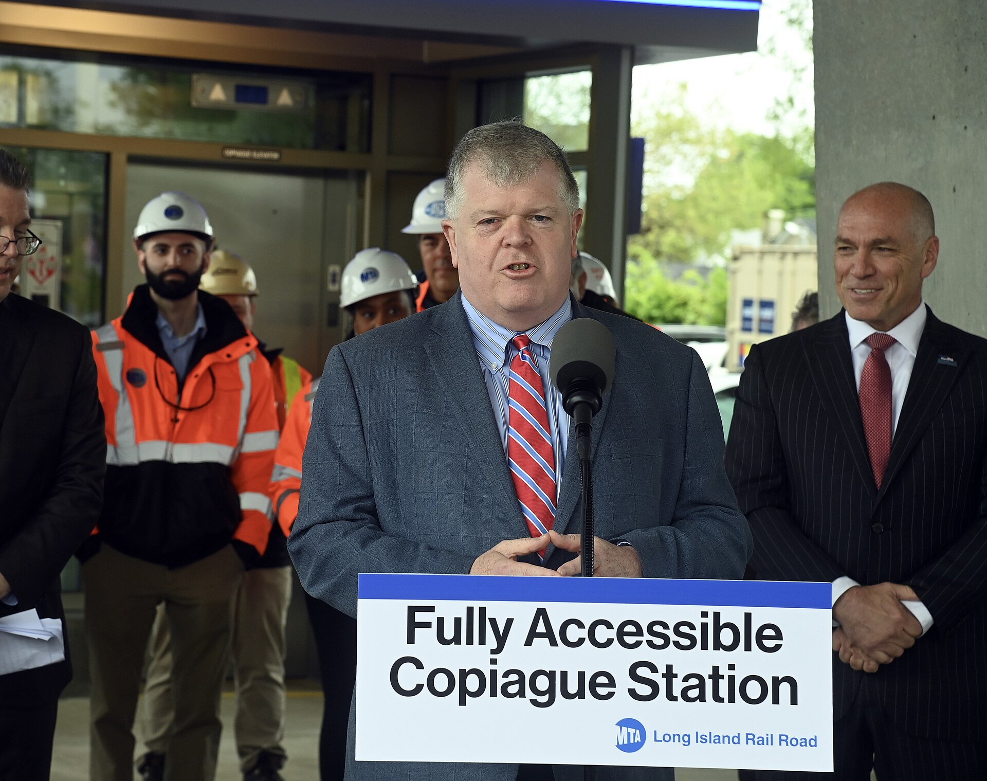 People stand beside the glass enclosure of a new station lift at Copiague railway station.