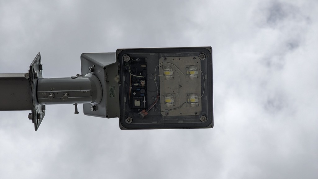 Underside of a rectangular LED street-light fitting mounted on a metal arm against a cloudy sky.