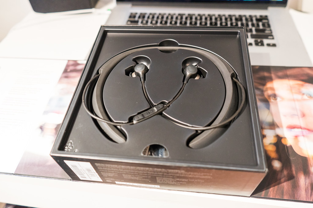 Black neckband-style Bose earphones resting in an open box, with a laptop behind them.